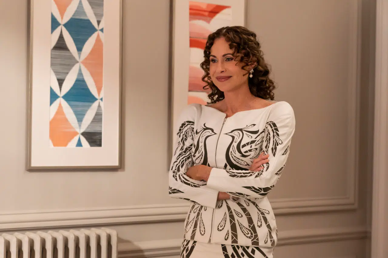 "Woman with curly hair in a patterned white outfit stands with arms crossed in a modern room with neutral walls, abstract art, and a radiator."