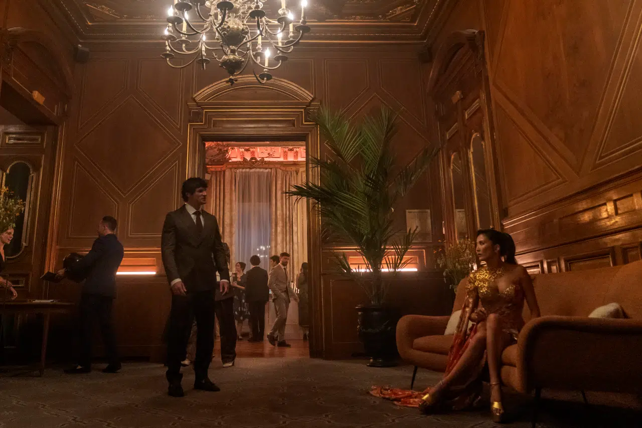 "A man in a suit stands in an elegant, wood-paneled lounge with a chandelier, while a woman in a gold dress sits on a couch; other people are visible in the background socializing at a formal event."
