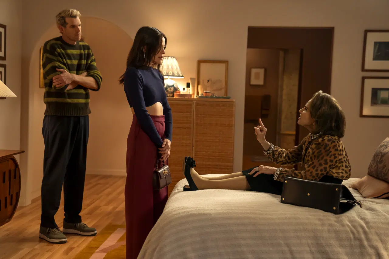 "Three people in a cozy, warmly lit bedroom; one woman sits on the bed gesturing, another stands with a purse, and a man in casual clothes stands nearby, watching the conversation."