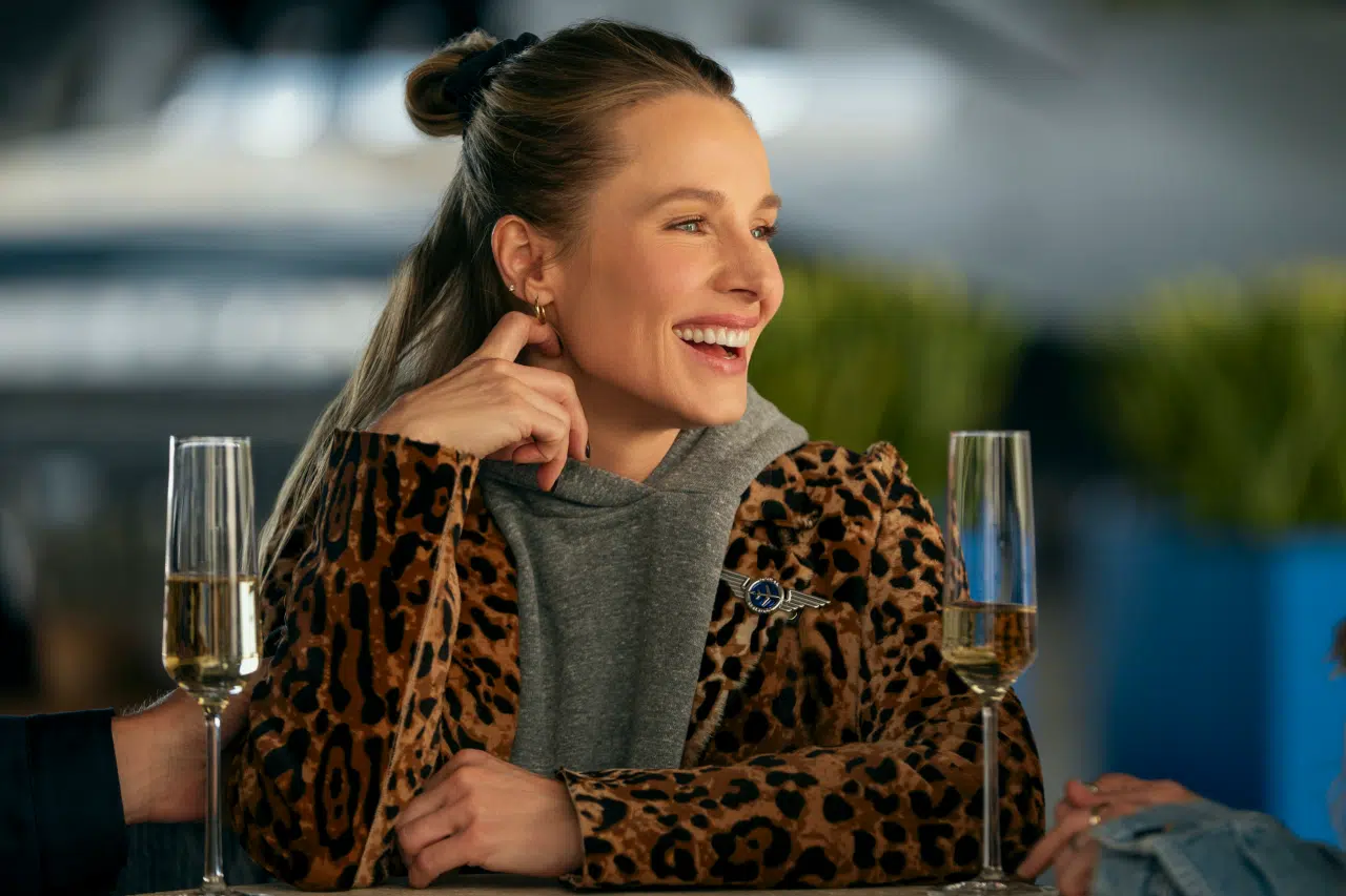 "Woman in leopard print jacket sitting at a table, smiling and talking with others, two glasses of champagne in front of her, modern indoor setting with blurred background and plants."