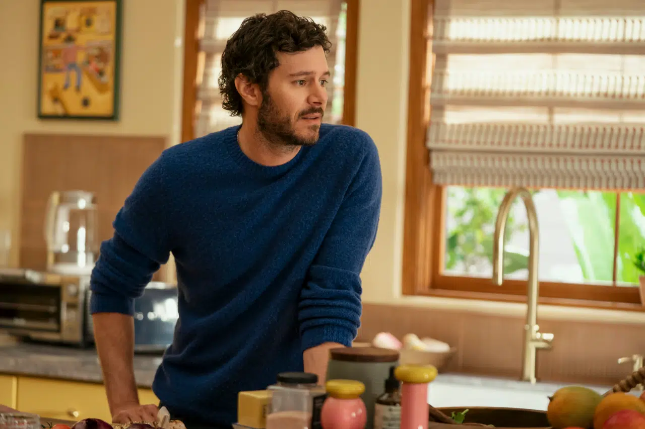 "A man with curly hair and a beard wearing a blue sweater stands in a cozy kitchen leaning on the counter, surrounded by cooking ingredients, with sunlight coming through the window blinds."