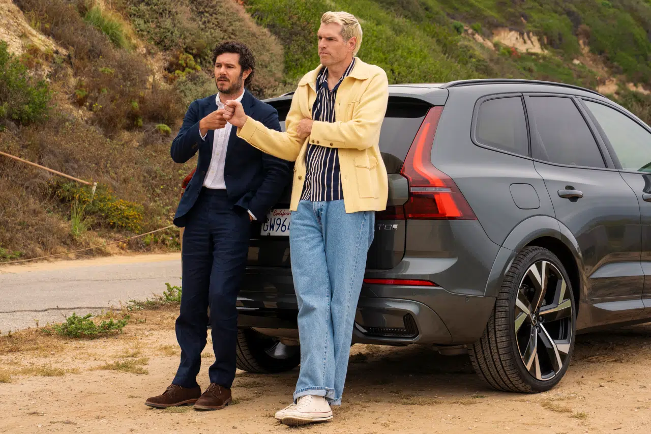 "Two men in casual and formal clothes stand outside, leaning against the back of a gray SUV parked on a dirt road, with green hills and vegetation in the background, appearing to have a conversation."