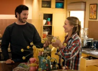 "A man and woman smile and toast with mugs in a cozy, sunlit kitchen, surrounded by yellow cabinets, kitchen appliances, and decorative items, creating a warm, homey atmosphere."