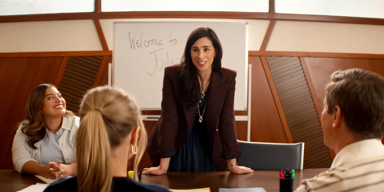 "A woman stands at the head of a conference table, smiling and addressing three colleagues in a meeting room. A whiteboard behind her reads "Welcome to" with another word partially visible."