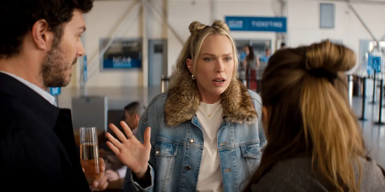 "Three people talking in an airport terminal, one woman in a jean jacket with fur collar gesturing animatedly, appearing to explain something to a man holding a drink and a woman with her back to the camera. Signs and check-in lines in background."