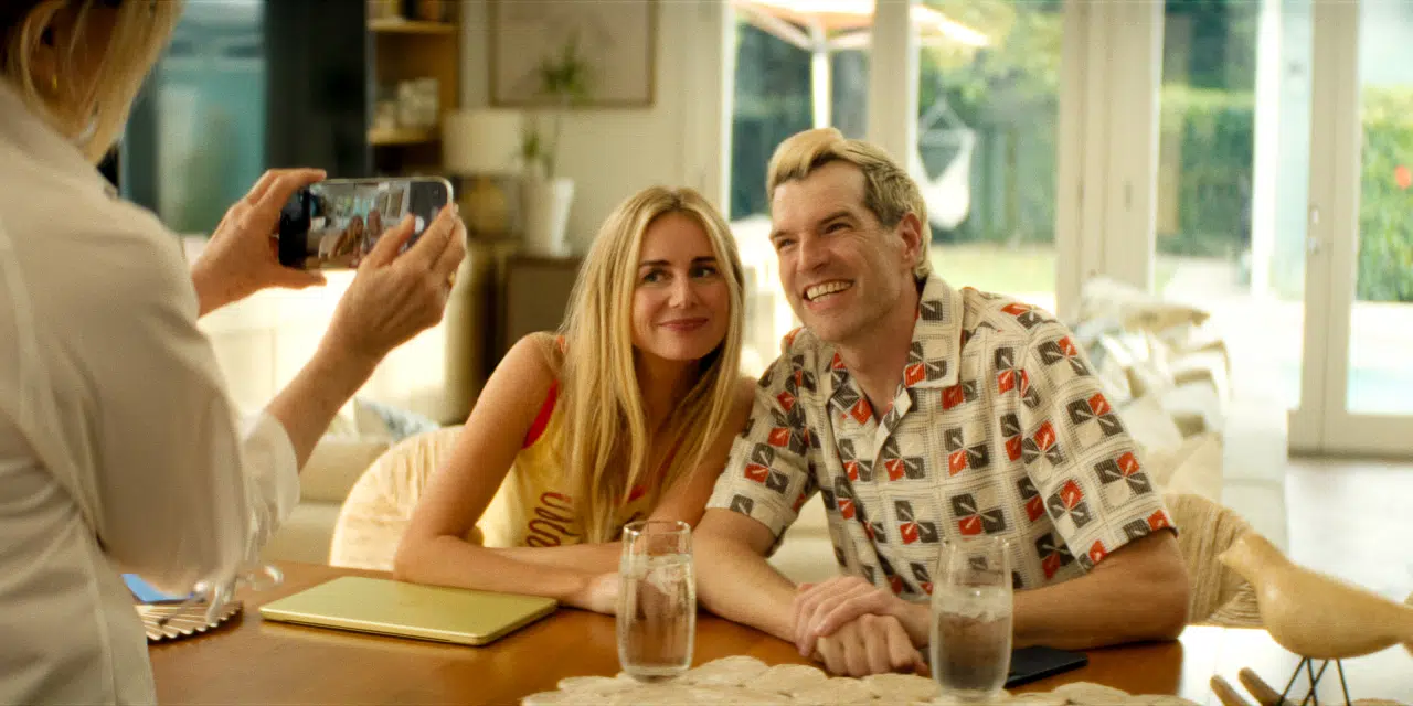 "A smiling couple sits at a table indoors, holding hands and posing for a photo taken by another person. Bright, cozy setting with large windows, plants outside, two glasses of water, and a yellow tablet on the table."
